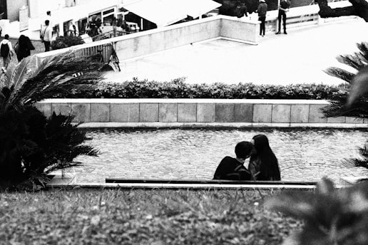 A black and white image captures a romantic moment between two people sitting closely on a bench, partially obscured by foliage in the foreground. The background shows a street scene with several pedestrians walking near a building.