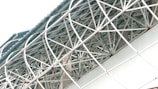 An intricate network of white metal beams forms a geometric pattern against a pale blue sky. The structure appears to be part of an architectural design, possibly a roof or a ceiling. The beams intersect at various angles, creating a lattice-like effect with red joint connectors visible at some of the intersections.