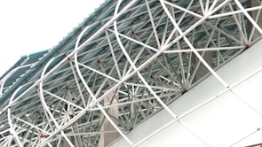 An intricate network of white metal beams forms a geometric pattern against a pale blue sky. The structure appears to be part of an architectural design, possibly a roof or a ceiling. The beams intersect at various angles, creating a lattice-like effect with red joint connectors visible at some of the intersections.