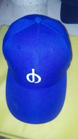 A blue baseball cap featuring a white embroidered symbol on the front panel. The cap is positioned on a light-colored surface with two other hats partially visible in the background.
