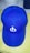 A blue baseball cap featuring a white embroidered symbol on the front panel. The cap is positioned on a light-colored surface with two other hats partially visible in the background.