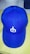 A blue baseball cap featuring a white embroidered symbol on the front panel. The cap is positioned on a light-colored surface with two other hats partially visible in the background.