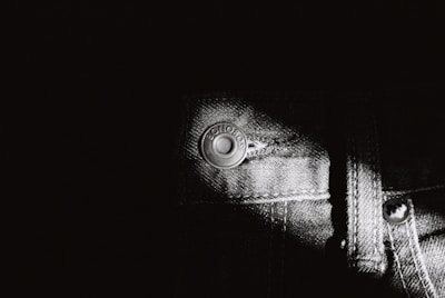 A close-up black and white photo of denim fabric showcasing a metal button with engraved text. The texture of the denim is highlighted with a strong contrast, emphasizing the weave and stitching details.