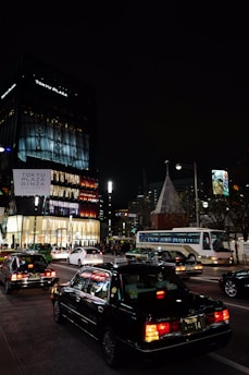 A bustling city street at night features several cars and taxis, with a large, illuminated shopping center labeled 'Tokyu Plaza' in the background. Bright lights and signage illuminate the scene, creating a lively urban atmosphere.