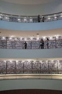 Several floors of a modern building interior feature images on walls, with people walking and viewing the displays. The architecture includes circular balconies with railings, and the walls are lined with many small, illuminated pictures. The setting appears quiet and contemplative, suggesting an exhibition or memorial space.