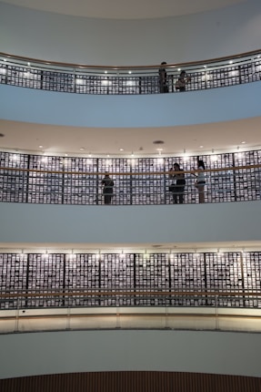 Several floors of a modern building interior feature images on walls, with people walking and viewing the displays. The architecture includes circular balconies with railings, and the walls are lined with many small, illuminated pictures. The setting appears quiet and contemplative, suggesting an exhibition or memorial space.