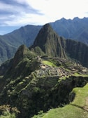 A panoramic view of the ancient Inca city of Machu Picchu, situated on a mountainous ridge with dramatic peaks and lush greenery surrounding the stone ruins. Terraced fields and stone structures are clearly visible, illustrating the historical and architectural significance of the site.