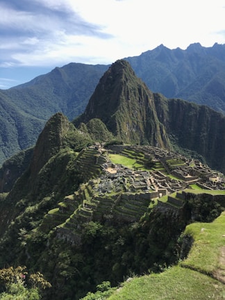 A panoramic view of the ancient Inca city of Machu Picchu, situated on a mountainous ridge with dramatic peaks and lush greenery surrounding the stone ruins. Terraced fields and stone structures are clearly visible, illustrating the historical and architectural significance of the site.