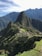 A panoramic view of the ancient Inca city of Machu Picchu, situated on a mountainous ridge with dramatic peaks and lush greenery surrounding the stone ruins. Terraced fields and stone structures are clearly visible, illustrating the historical and architectural significance of the site.