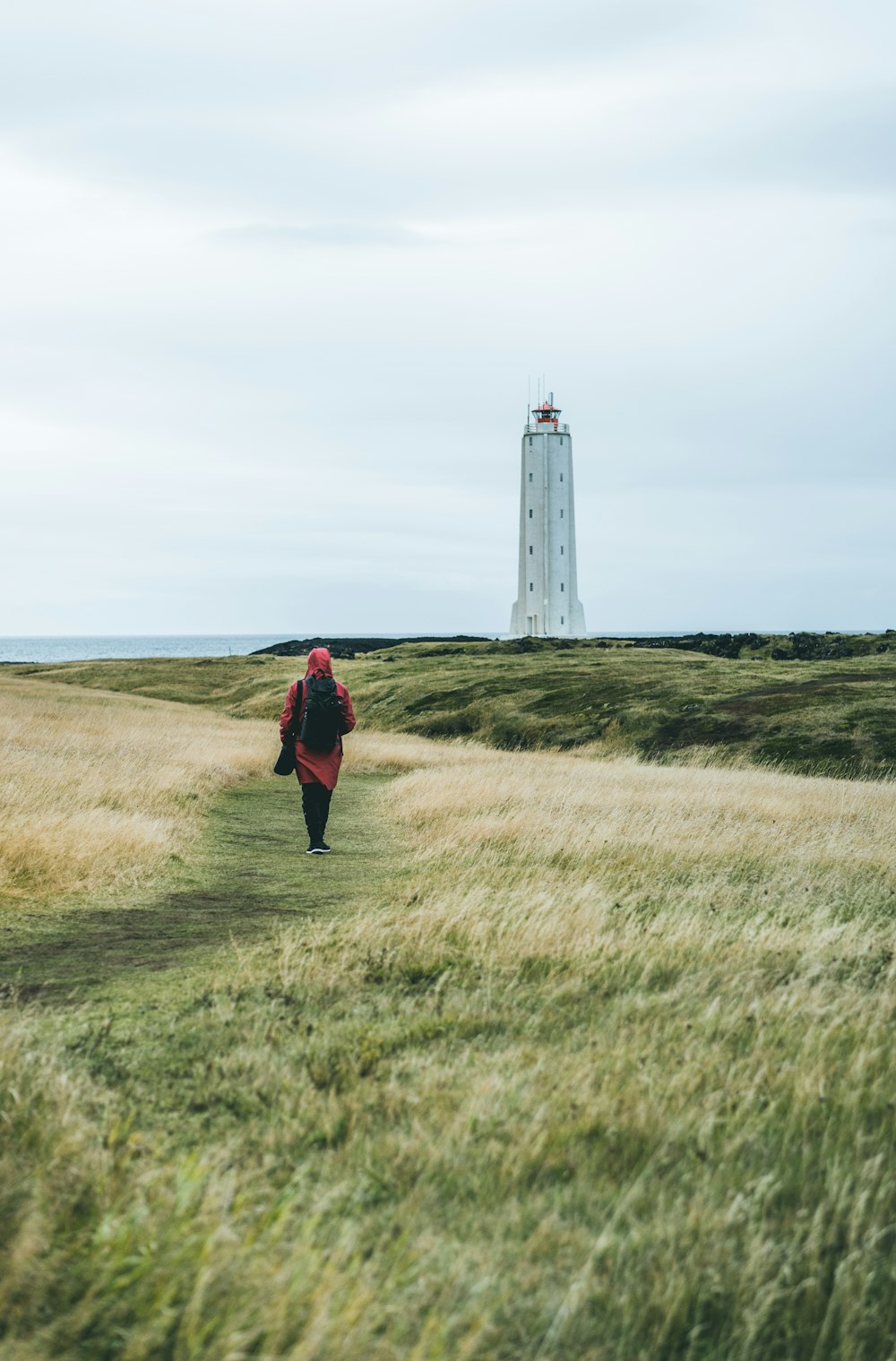 femme en robe rouge marche sur l’herbe verte menant à un phare blanc