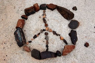 An arrangement of rocks forms the shape of a peace symbol on a sandy surface. The rocks vary in size and color, including dark brown, reddish, and black tones. The peace symbol is neatly composed, contrasting with the light-colored sandy background.