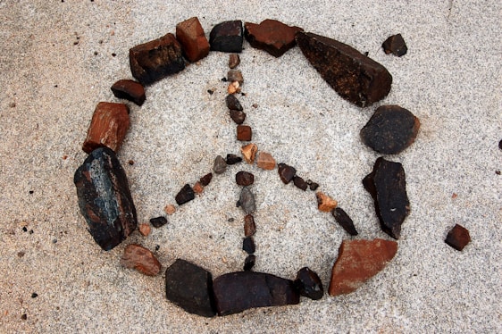 An arrangement of rocks forms the shape of a peace symbol on a sandy surface. The rocks vary in size and color, including dark brown, reddish, and black tones. The peace symbol is neatly composed, contrasting with the light-colored sandy background.