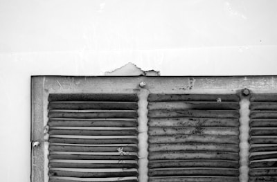 A weathered and rusty metal vent with horizontal slats is attached to a wall. The paint on the wall is peeling above the vent, and there are small signs of deterioration. The vent appears aged, with visible grime and scratches on its surface.