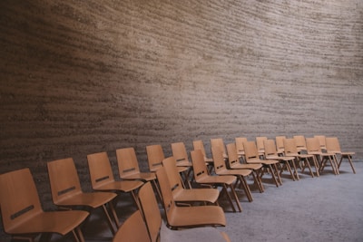 A series of simple wooden chairs arranged in parallel rows within a minimalist room with textured concrete walls.