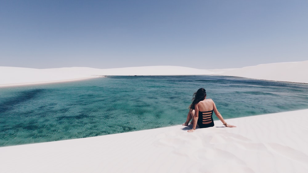 woman sitting on white sand near body of water