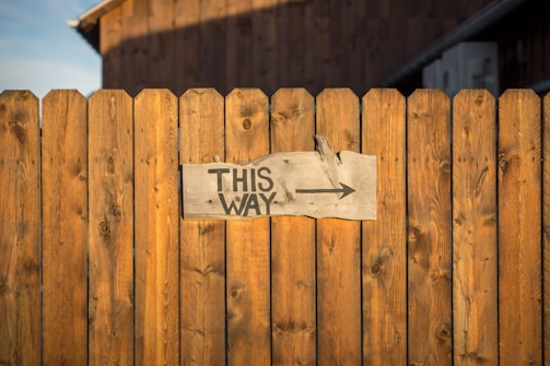 A wooden fence with a weathered, rectangular sign attached. The sign has bold black text reading 'THIS WAY' and an arrow pointing to the right. The fence is made of vertical wooden planks with a warm brown hue. The background shows a partially visible wooden structure.