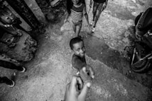 A black and white photograph capturing a joyful child reaching out towards the camera with a big smile. Another child and an adult are partially visible in the background. The setting appears to be an outdoor urban environment with rough textured ground and scattered objects.