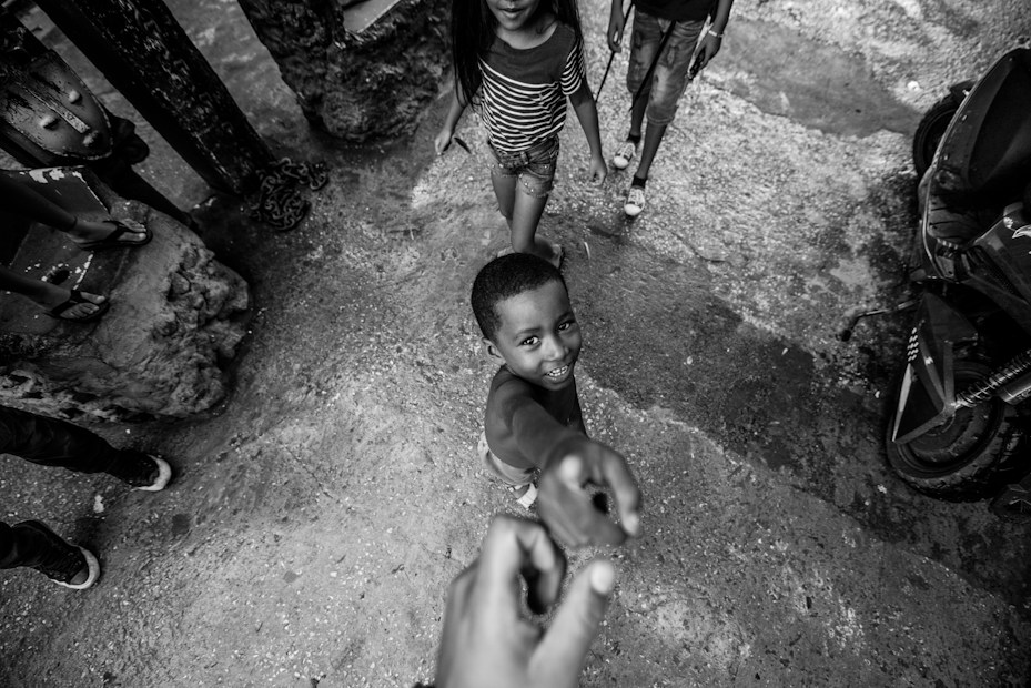 A black and white photograph capturing a joyful child reaching out towards the camera with a big smile. Another child and an adult are partially visible in the background. The setting appears to be an outdoor urban environment with rough textured ground and scattered objects.