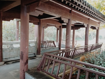 A traditional wooden pavilion with intricate railings and a tiled roof, set in a natural environment with lush greenery surrounding it. The structure features multiple posts supporting the roof, and pathways leading through the pavilion. The scene appears tranquil and serene.