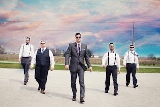 Five men are walking confidently on a road under a vibrant, colorful sky. The man in the center wears a grey suit and sunglasses, while the others are dressed in suspenders and ties. The background shows open fields and a few buildings in the distance.