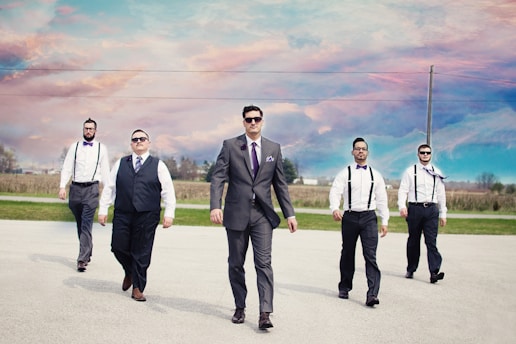 Five men are walking confidently on a road under a vibrant, colorful sky. The man in the center wears a grey suit and sunglasses, while the others are dressed in suspenders and ties. The background shows open fields and a few buildings in the distance.