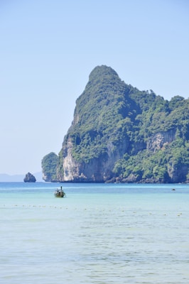 A serene coastal scene with a small traditional boat floating on clear blue waters. The backdrop features a large, lush green rocky island with steep cliffs rising dramatically from the sea, covered in dense foliage. The sky is bright and clear, enhancing the tranquil and picturesque setting.
