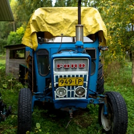 A vintage blue Ford tractor with a yellow tarp covering the top is parked on green grass. The tractor has a prominent grille with the brand name in red letters and a license plate numbered 949-IP. Surrounding greenery includes trees and shrubs, creating a rustic outdoor setting.