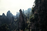 Tall, narrow rock formations covered with lush greenery rise dramatically against a hazy sky. The landscape appears untouched and mystical, with the vertical cliffs emerging like natural towers from dense forests below.