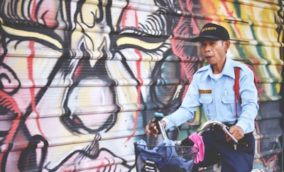 A man in a security uniform rides a bicycle past a vibrantly colored graffiti mural on a wall. His uniform consists of a light blue shirt with a security badge and a matching cap. The mural features abstract designs with bold lines and a mix of colors like red, yellow, and black.