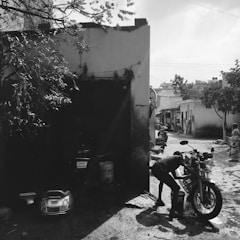 A person is washing a motorcycle in a narrow alleyway beside a small building. There are buckets and a detached motorbike seat on the ground. The area is shaded by trees, and other motorcycles are parked further down the alley. A woman in traditional clothing stands near the entrance of a building. The scene is in black and white, adding a nostalgic and timeless feel.