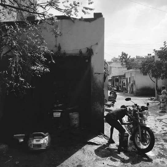 A person is washing a motorcycle in a narrow alleyway beside a small building. There are buckets and a detached motorbike seat on the ground. The area is shaded by trees, and other motorcycles are parked further down the alley. A woman in traditional clothing stands near the entrance of a building. The scene is in black and white, adding a nostalgic and timeless feel.