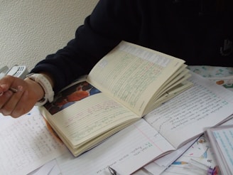 A person with a watch on their wrist is holding a pen and writing on a notebook. There are multiple sheets of paper around the notebook, filled with handwritten text. The scene suggests a study or work environment, with attention focused on writing.
