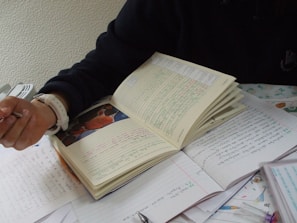 A person with a watch on their wrist is holding a pen and writing on a notebook. There are multiple sheets of paper around the notebook, filled with handwritten text. The scene suggests a study or work environment, with attention focused on writing.