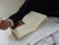 A person with a watch on their wrist is holding a pen and writing on a notebook. There are multiple sheets of paper around the notebook, filled with handwritten text. The scene suggests a study or work environment, with attention focused on writing.