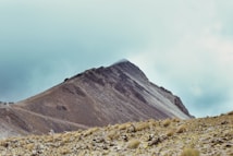 A rugged mountain slope with a sharp peak is under a cloudy sky. The terrain features sparse vegetation, predominantly composed of dry grass and rocks. A line of small figures can be seen ascending the slope, indicating a group of hikers. The scene evokes a sense of isolation and adventure in a natural, high-altitude setting.
