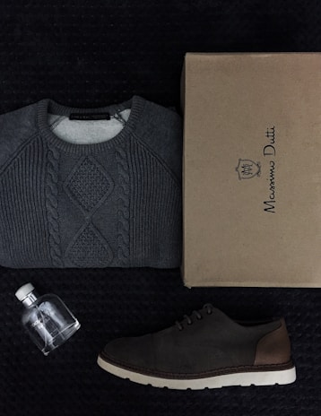 A neatly folded gray cable-knit sweater is placed alongside a brown shoe and a cardboard box labeled Massimo Dutti. There is also a small, clear glass bottle with a white cap, likely a perfume, positioned nearby. All items are arranged on a textured dark surface.
