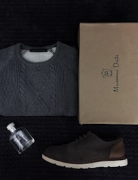 A neatly folded gray cable-knit sweater is placed alongside a brown shoe and a cardboard box labeled Massimo Dutti. There is also a small, clear glass bottle with a white cap, likely a perfume, positioned nearby. All items are arranged on a textured dark surface.