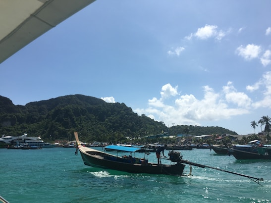 A traditional long-tail boat is floating on a vibrant blue-green sea, with lush green mountains in the background under a clear blue sky with a few fluffy clouds. Other boats and structures can be seen along the shoreline, creating a picturesque and serene coastal scene.