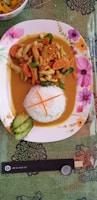 A colorful plate of food featuring rice molded into a cylindrical shape, partially covered in a golden-orange curry with pieces of chicken, carrots, and green vegetables. A few slices of cucumber are arranged beside the rice, and the plate is decorated with pink and yellow floral patterns. The background is a green patterned tablecloth, and there is a pair of chopsticks and a bottle cap nearby.