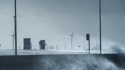 Wind turbines are visible in the distance on a stormy day, with splashing waves hitting a concrete walkway. Two lampposts and railings are also present along the path, creating a sense of infrastructure facing the turbulent sea.