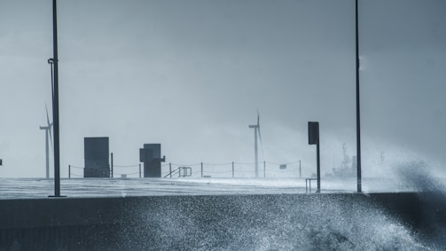 Wind turbines are visible in the distance on a stormy day, with splashing waves hitting a concrete walkway. Two lampposts and railings are also present along the path, creating a sense of infrastructure facing the turbulent sea.