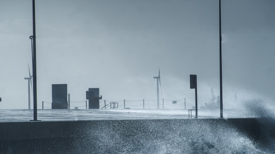 Wind turbines are visible in the distance on a stormy day, with splashing waves hitting a concrete walkway. Two lampposts and railings are also present along the path, creating a sense of infrastructure facing the turbulent sea.