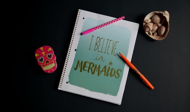 A spiral notebook with a cover reading 'I Believe in Mermaids' in gold writing on a light blue background. Surrounding the notebook are a pink pen with a zigzag pattern, an orange pen with a floral design, a decorative skull-shaped tin with colorful hearts and flowers, and a seashell containing smaller shells.