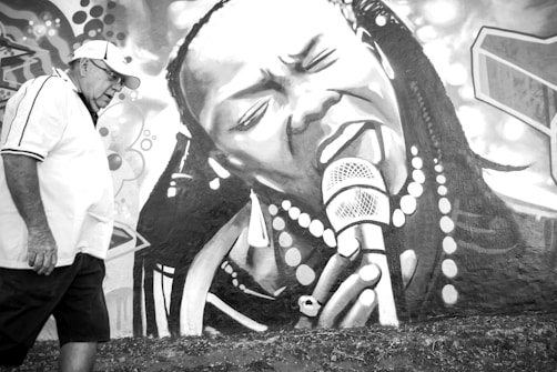 A black and white mural depicts a woman passionately singing into a microphone, adorned with large round beads and earrings. A man in a white shirt and cap walks past, glancing at the artwork. The mural fills the entire wall with abstract elements in the background.