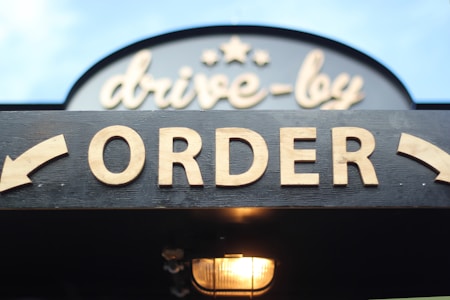 A sign for a drive-through ordering area, featuring bold wooden letters spelling 'ORDER' against a dark background, with an arrow pointing to the left. Above this, a decorative sign displays 'drive-by' with stars, and a light fixture is visible below.