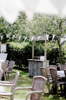 A serene outdoor setting featuring wooden tables and chairs arranged on a grassy lawn. A rustic bar structure with glasses is present in the background, adorned with a 'Mr & Mrs' sign. White triangular bunting is strung between trees, casting gentle shadows. The lush greenery and trees in the background evoke a peaceful, natural atmosphere.