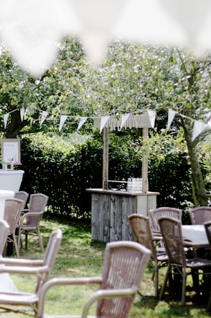 A serene outdoor setting featuring wooden tables and chairs arranged on a grassy lawn. A rustic bar structure with glasses is present in the background, adorned with a 'Mr & Mrs' sign. White triangular bunting is strung between trees, casting gentle shadows. The lush greenery and trees in the background evoke a peaceful, natural atmosphere.