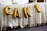 Gold foil balloons spelling out the word 'CAKE' are displayed against a lace tablecloth. The table is covered with decorative food items and containers on top.