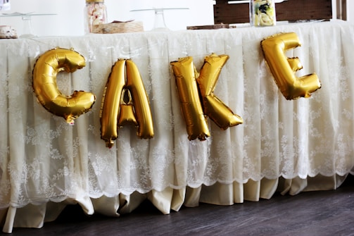 Gold foil balloons spelling out the word 'CAKE' are displayed against a lace tablecloth. The table is covered with decorative food items and containers on top.