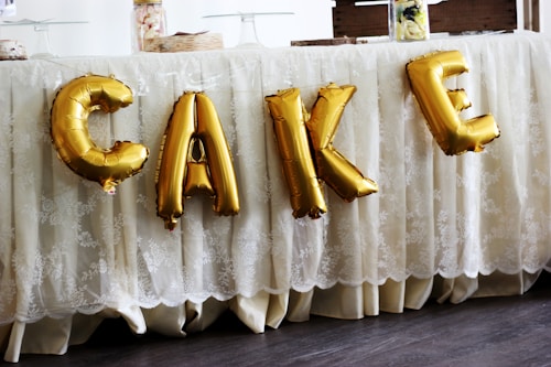 Gold foil balloons spelling out the word 'CAKE' are displayed against a lace tablecloth. The table is covered with decorative food items and containers on top.